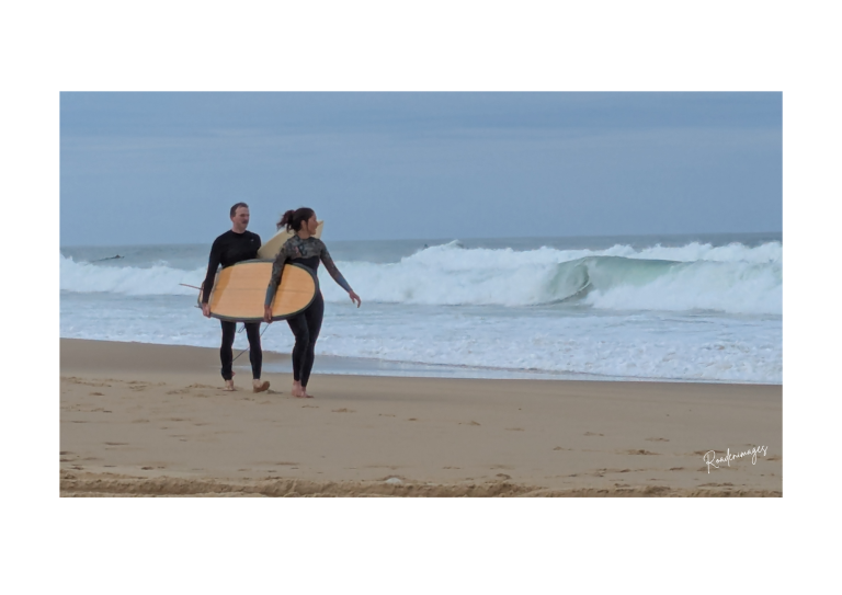 Longboard in love Deux surfeurs marchent sur la plage avec une planche de surf, vagues en arrière-plan.