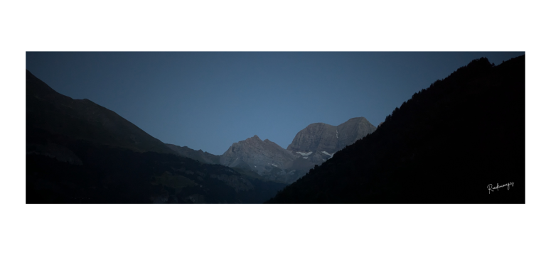 Montagnes sombres sous un ciel bleu crépusculaire, avec des sommets enneigés au loin.